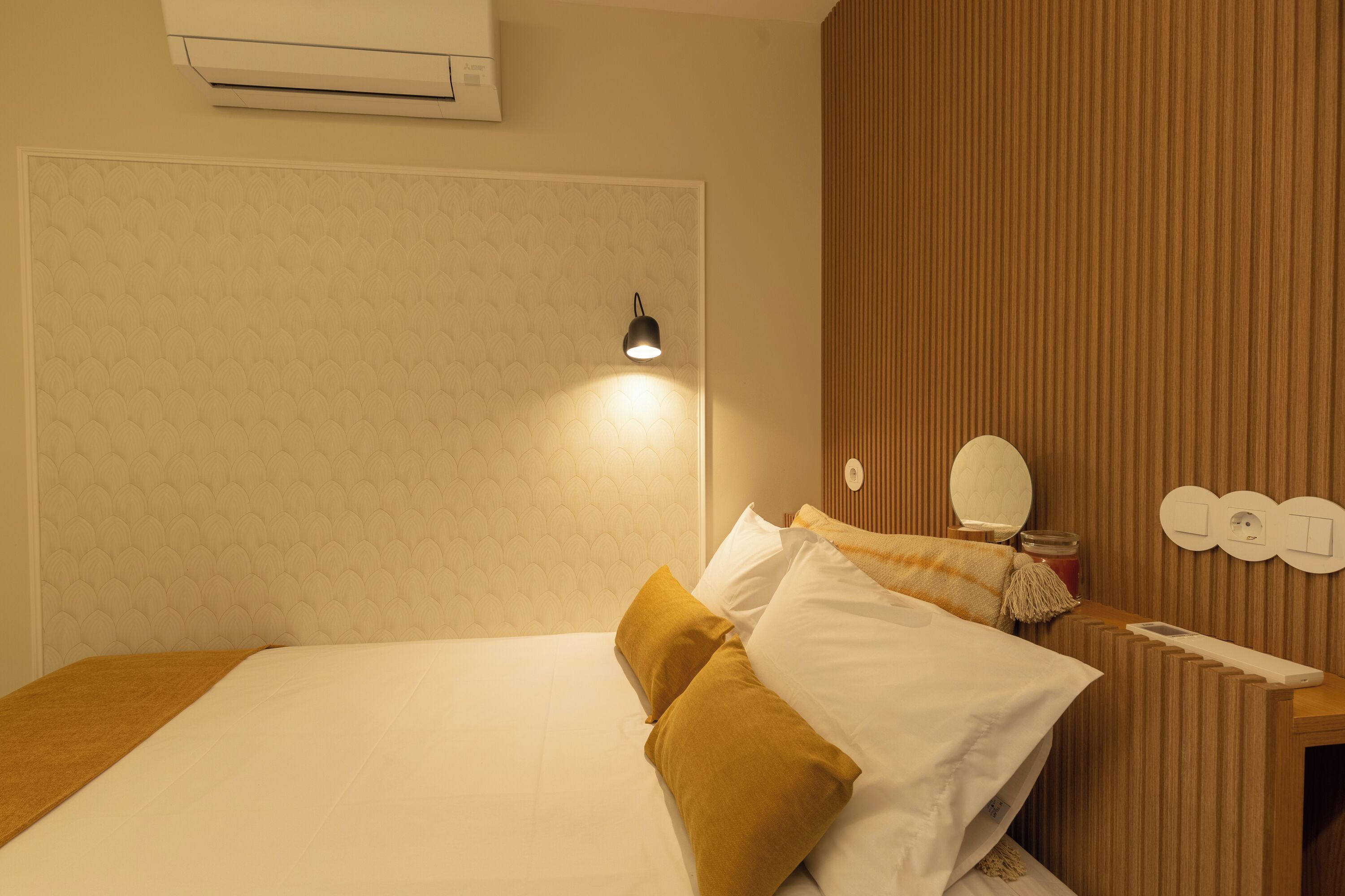 A hotel room with a bed, a bedside table with a lamp, a mirror, and a wall-mounted air conditioner.