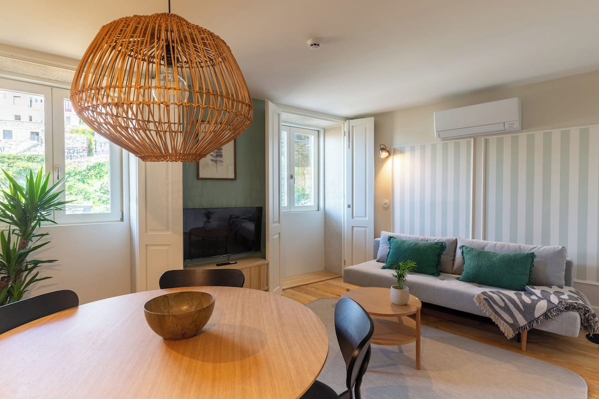 A modern living room with a round wooden dining table, a woven pendant light, a sofa with green cushions, and a television on a stand.