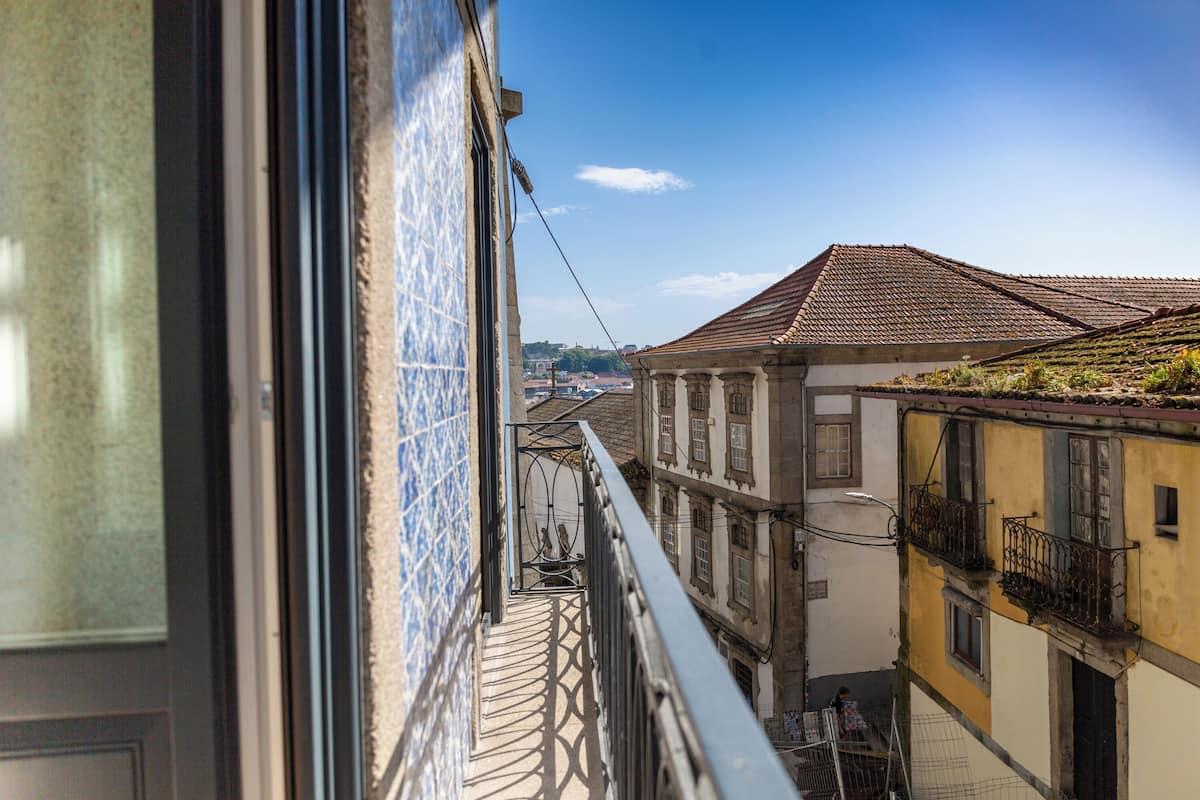 A narrow street with buildings on either side, a balcony with a metal railing, and a clear blue sky.