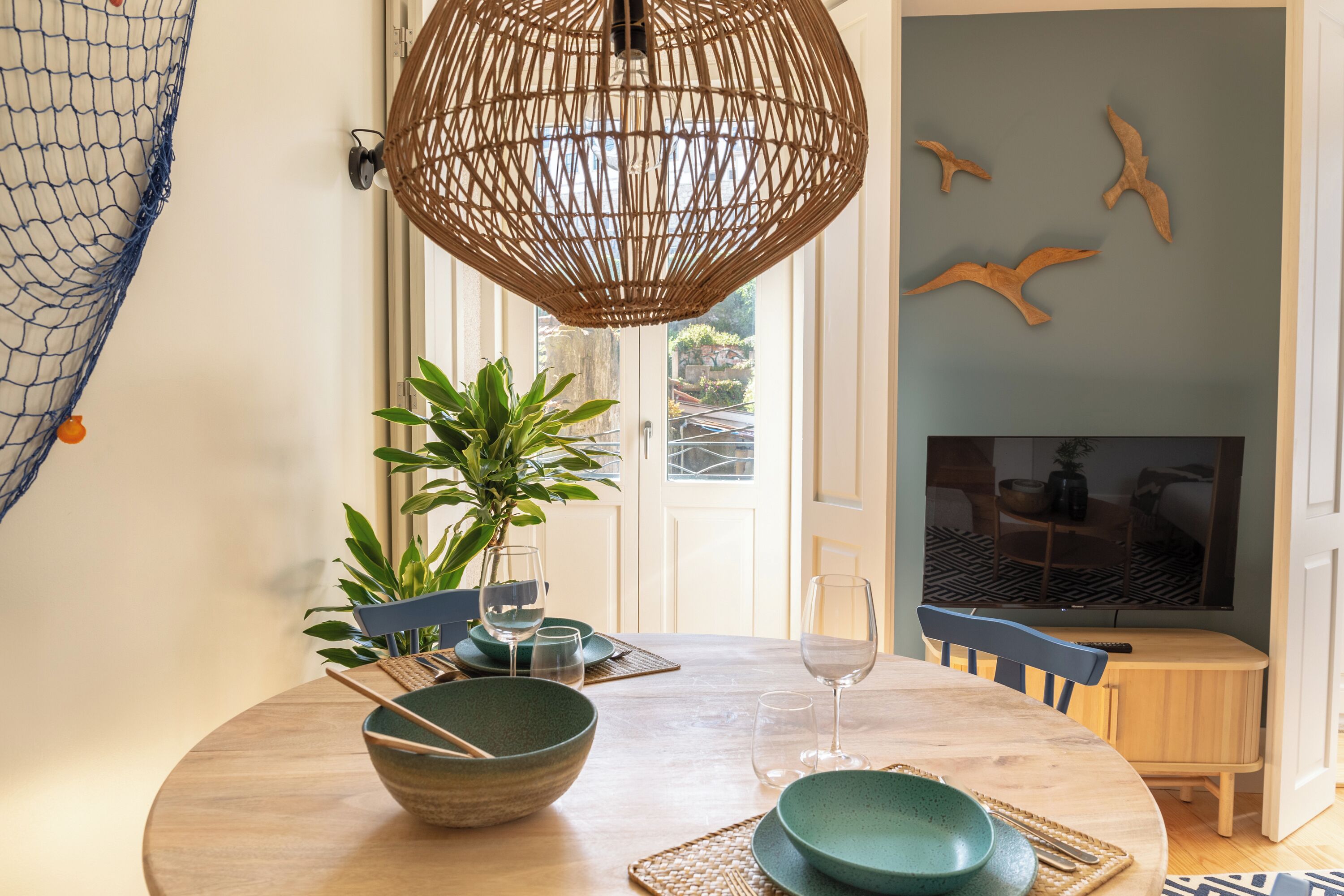 Et spiseområde med et rundt bord, stole, en vævet lampe, en potteplante og et fjernsyn i baggrunden.
