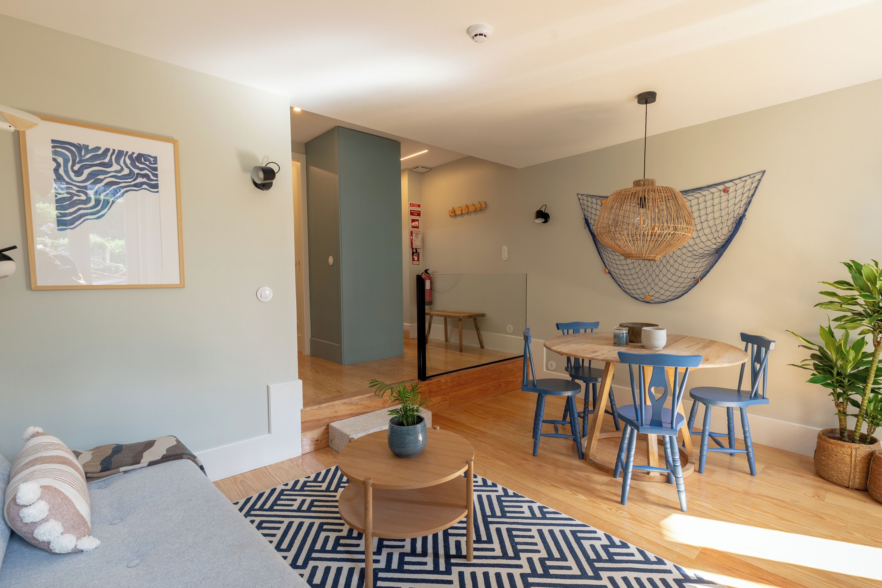 Un salon moderne avec un plancher en bois, un tapis à motifs bleus, un coin repas avec des chaises bleues, un luminaire suspendu et une œuvre d’art encadrée au mur.