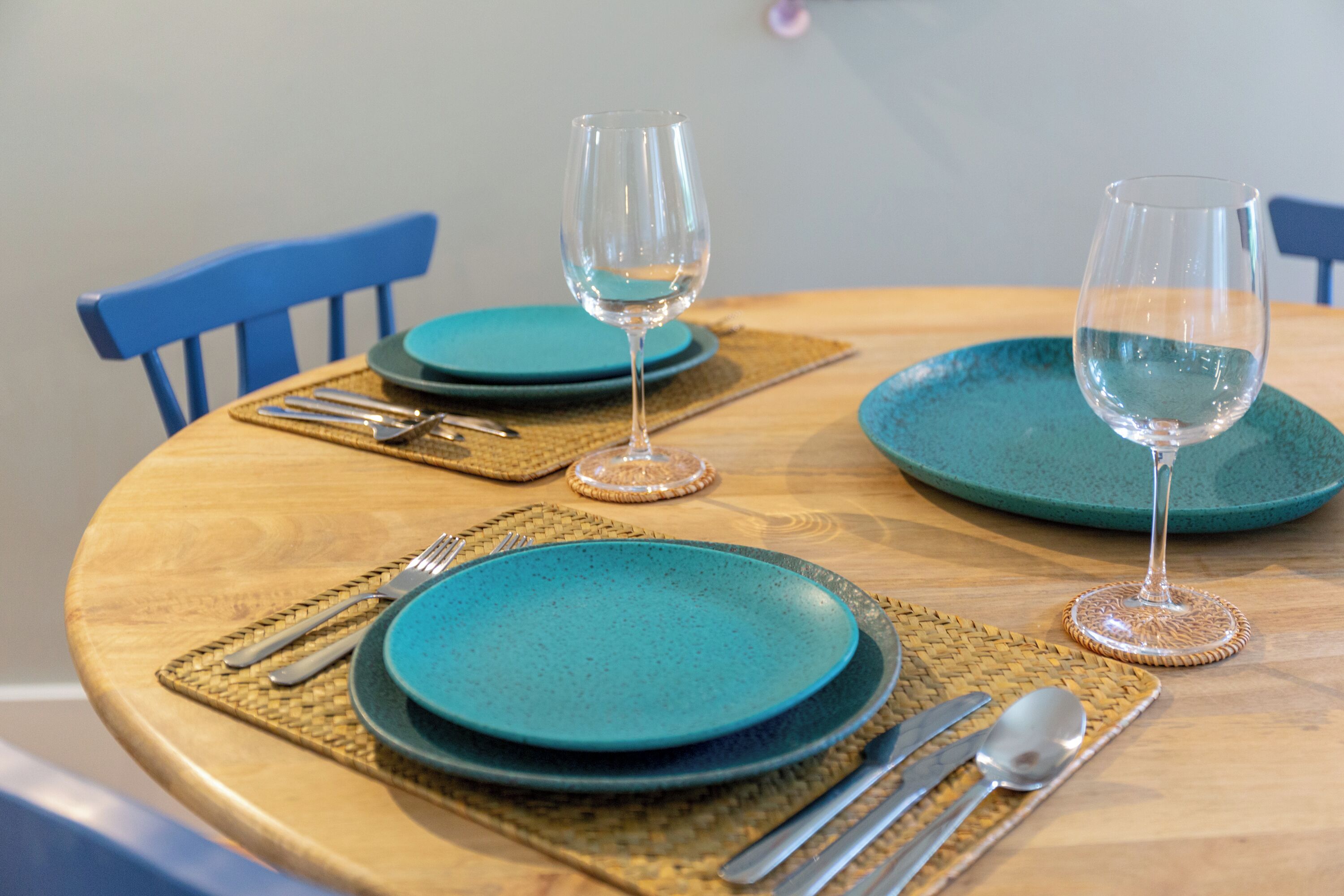 A round wooden table with teal plates, cutlery, and two wine glasses.