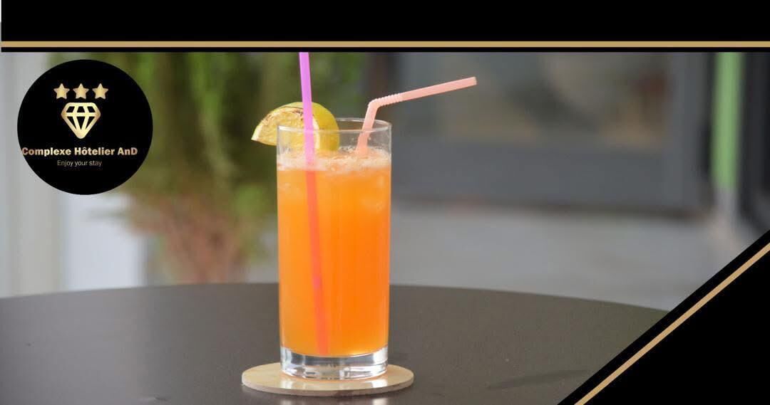 A tall glass with an orange beverage, garnished with a lemon slice, and two straws, placed on a round table.