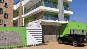 Un edificio moderno de varios pisos con fachada verde y blanca, una escalera central y un SUV negro estacionado en frente.