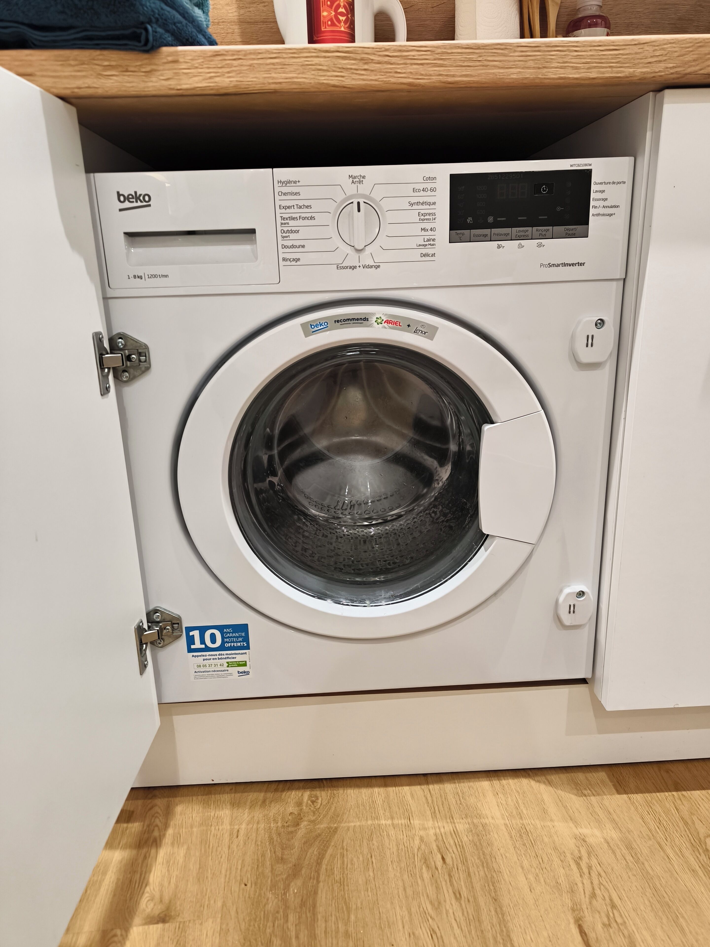 Una lavadora de carga frontal con un panel de control, una etiqueta de garantía de 10 años y un estante de madera encima.