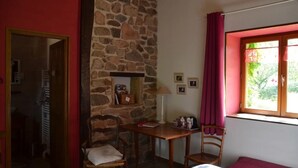 Ein Zimmer mit Steinwand, einem Holzstuhl, einem Tisch mit Lampe, einem Bett mit rotem Vorhang und einem Fenster mit Blick auf Grün.