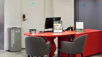 A reception desk with a red table and two grey chairs, a computer, and a sign that reads 'Bonjour & Bienvenue' with 'Hello & welcome' in English.