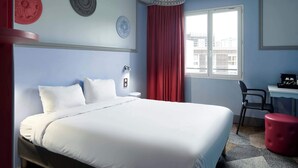 A hotel room with a bed, a desk, a chair, a red curtain, and a window with a city view. - B&B Hotel Saint-maur Créteil (Saint-Maur-des-Fosses)