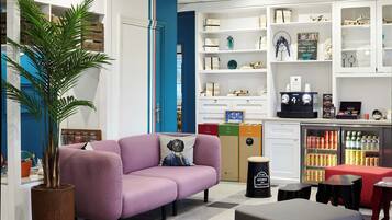 Een modern interieur met een paarse bank, een grote potplant, een koffieapparaat en een koelkast vol drankjes.