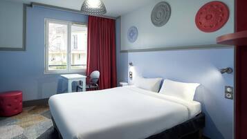 Een hotelkamer met een bed, een bureau, een stoel, een rode poef en een raam met gordijnen.