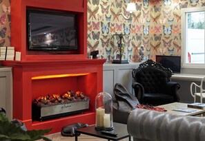 A living room with a red fireplace, a flat-screen TV, a patterned wallpaper, a black armchair, and a white chair.