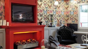A living room with a red fireplace, a flat-screen TV, a patterned wallpaper, a black armchair, and a white chair. - B&B Hotel Saint-maur Créteil (Saint-Maur-des-Fosses)