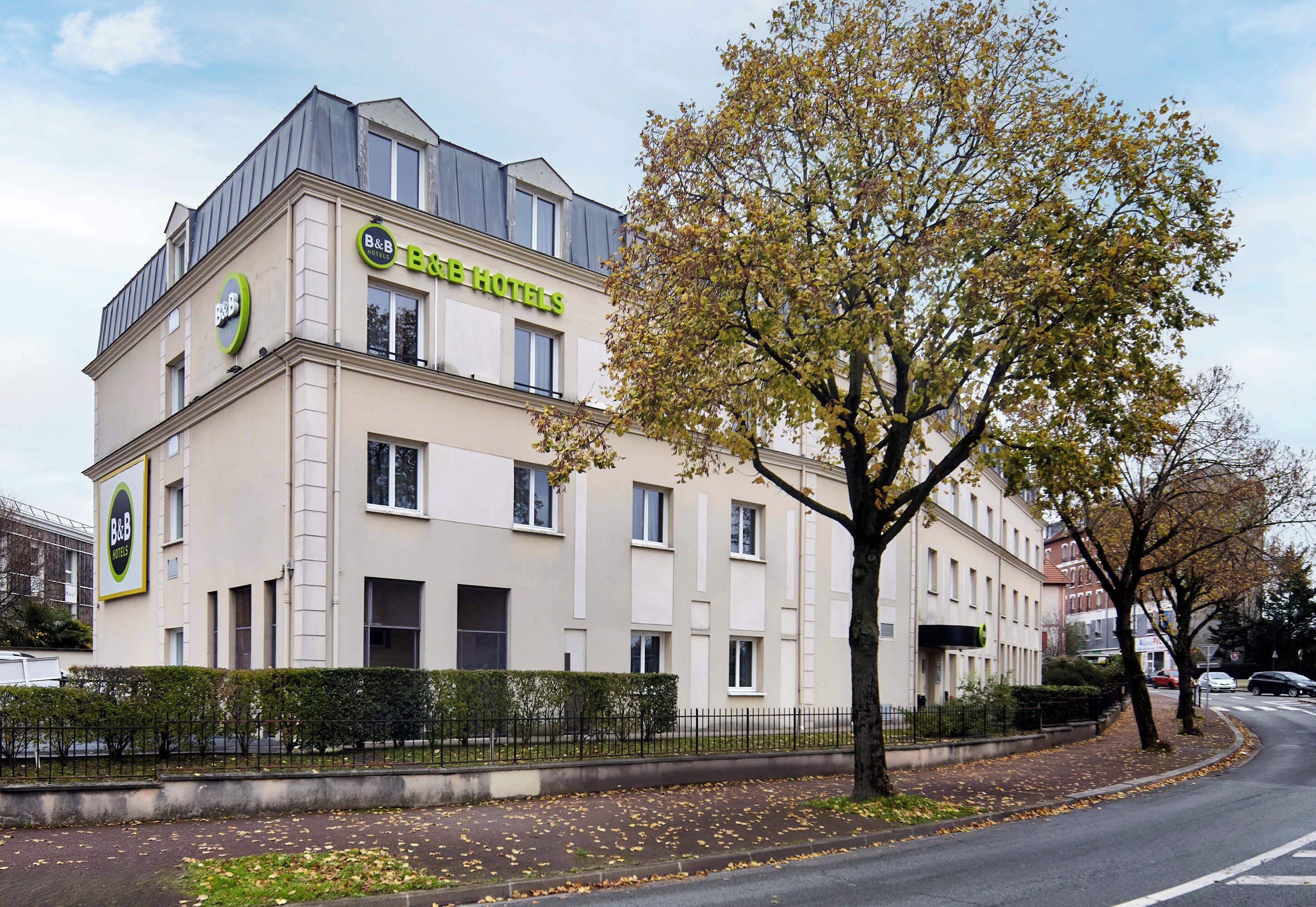 A B&B hotel with a modern design, a prominent B&B logo, and a tree in front.