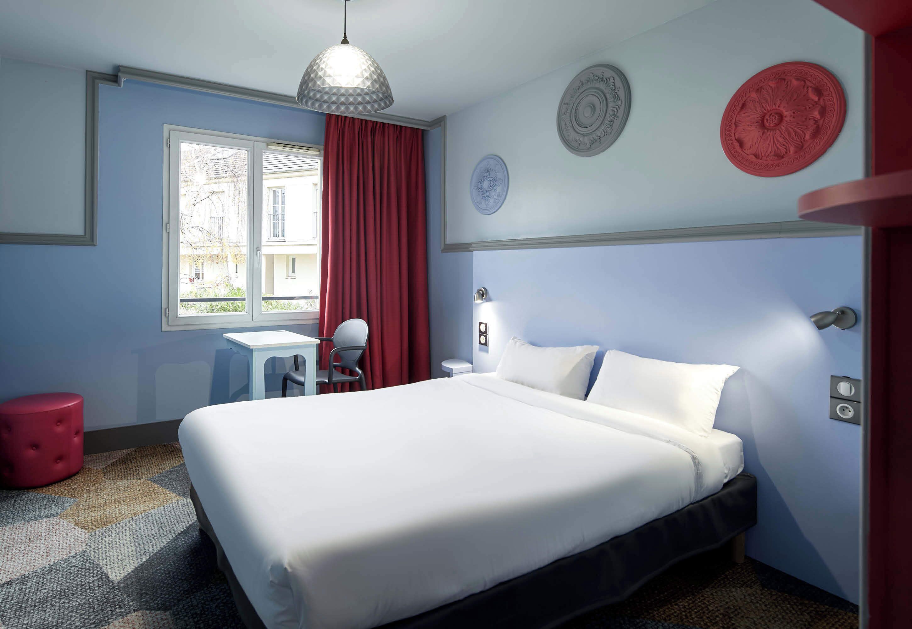 A hotel room with a bed, a desk, a chair, a red ottoman, and a window with curtains.