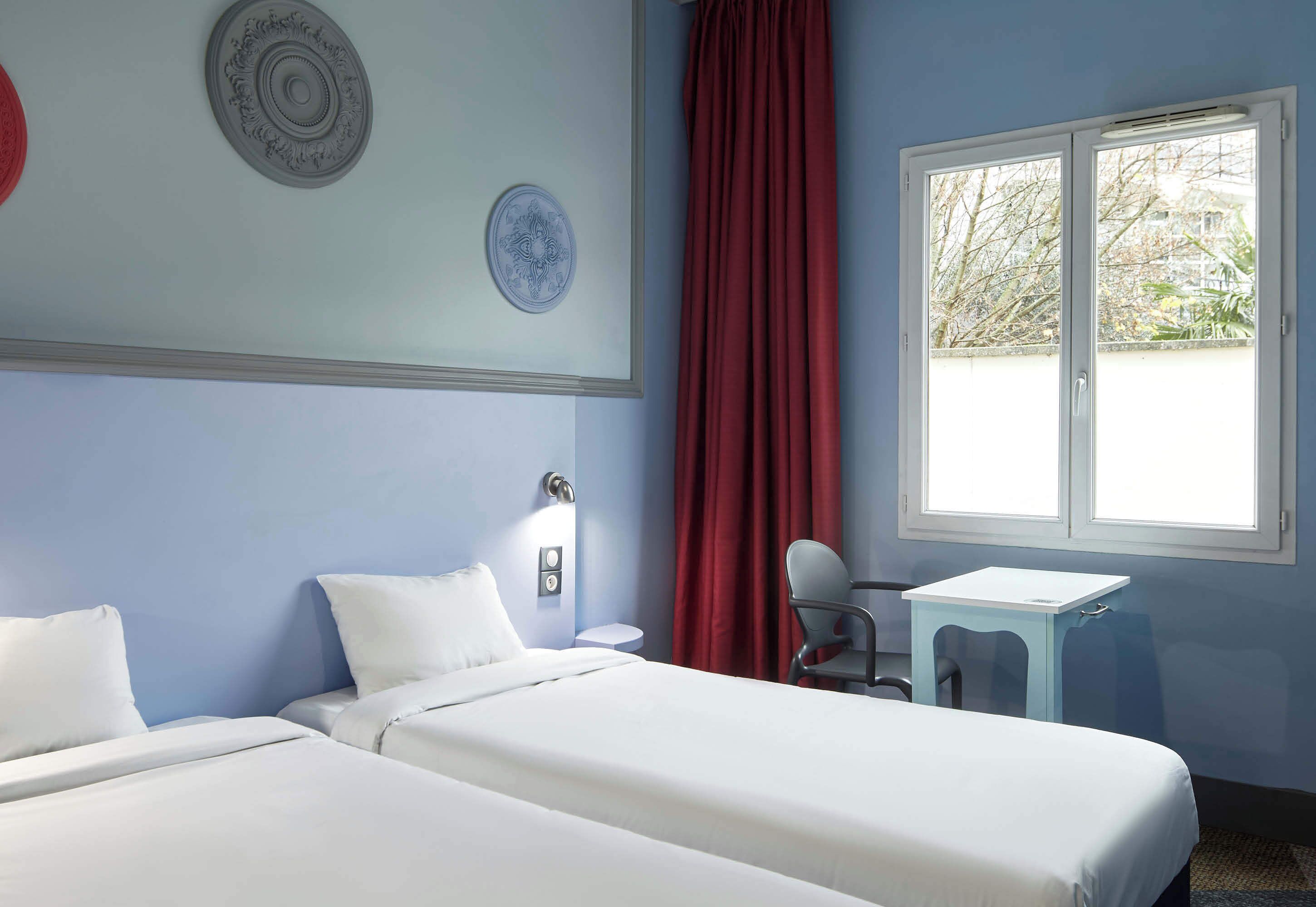 A room with two beds, a desk, a chair, a window with curtains, and a wall-mounted light fixture.