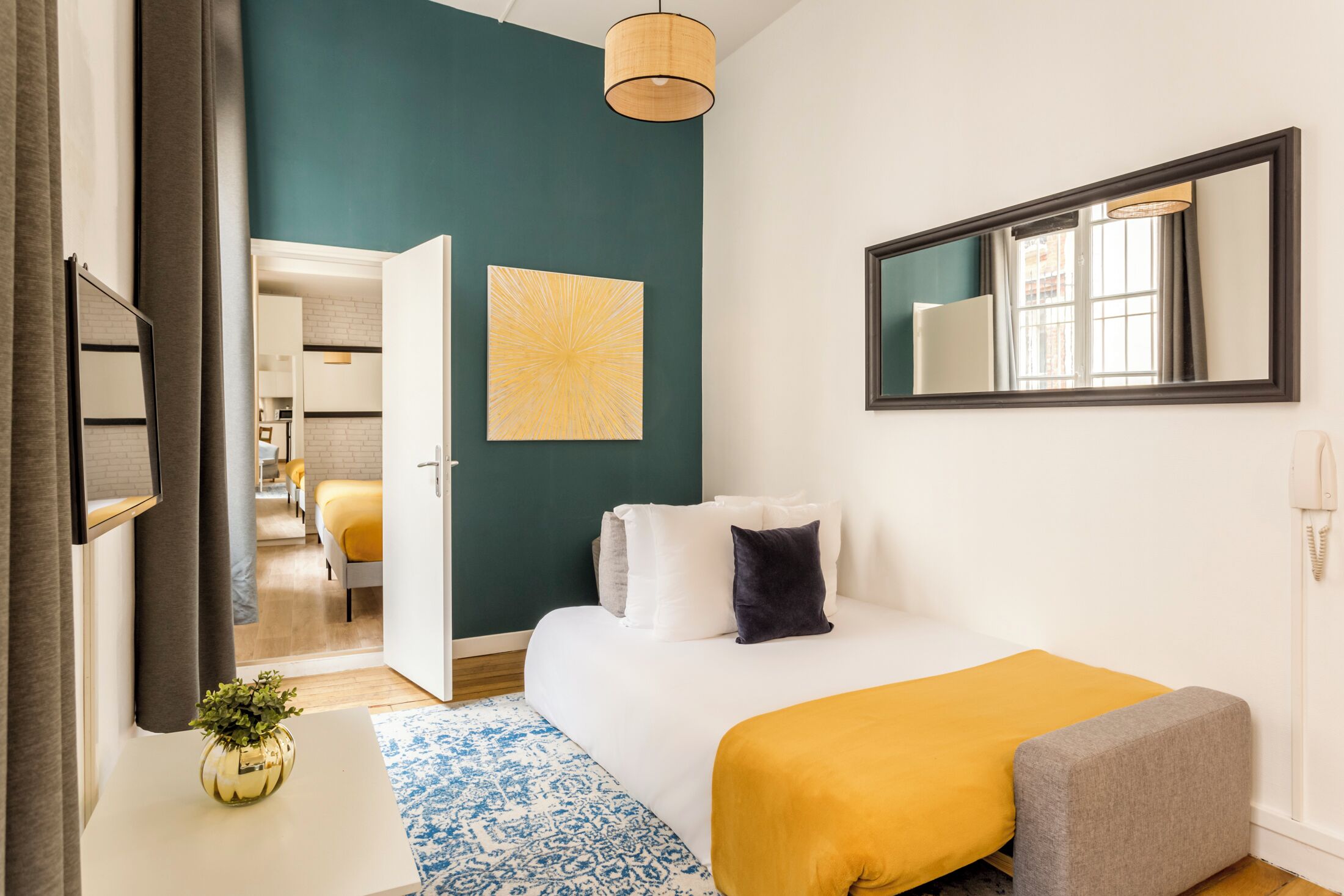 Une chambre moderne avec un lit, une table de chevet agrémentée d’une plante, un grand miroir et une vue sur la pièce adjacente.