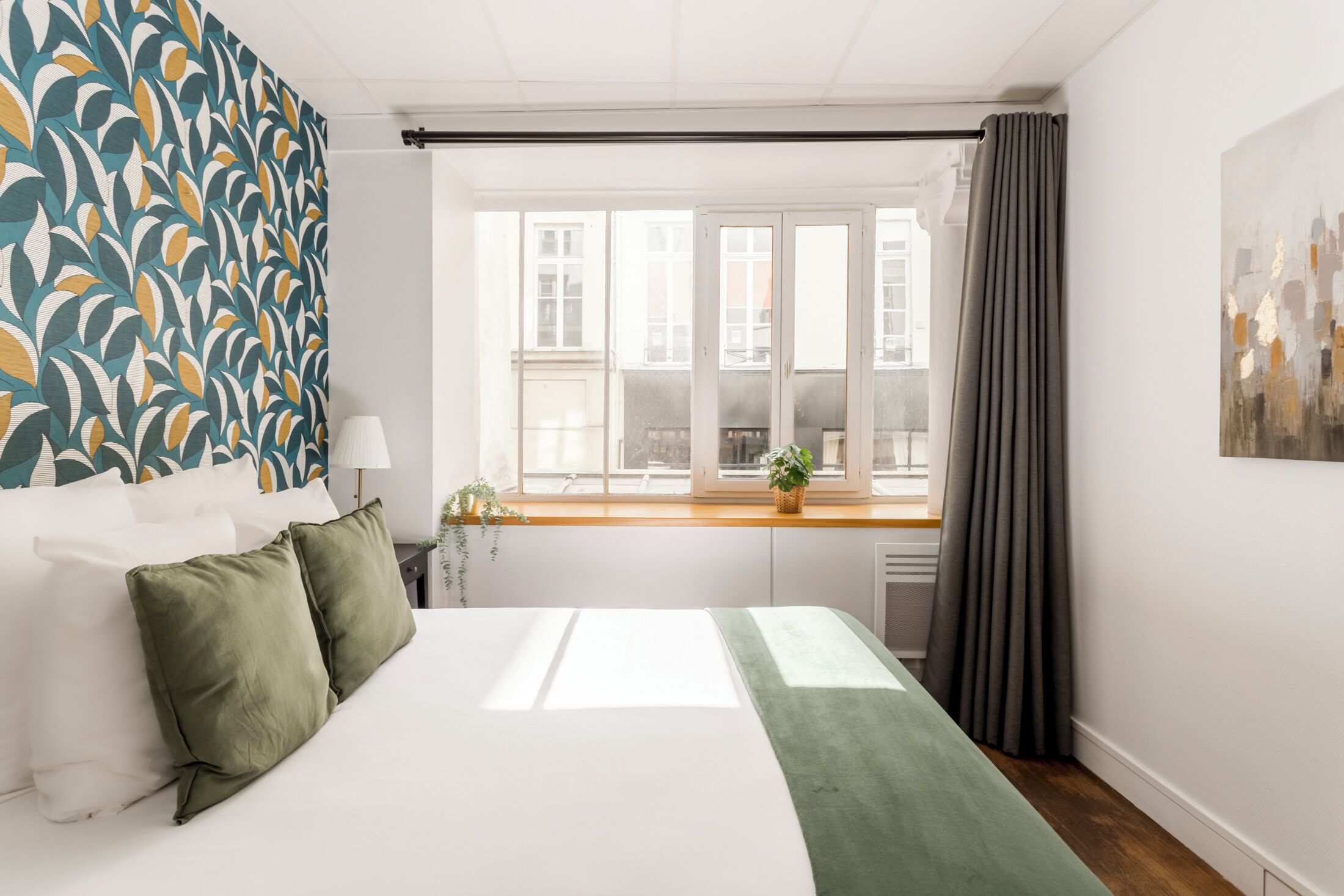 A bedroom with a large bed, a window with curtains, a view of buildings outside, and a patterned wall.