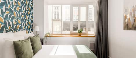 A bedroom with a large bed, a window with curtains, a view of buildings outside, and a patterned wall.