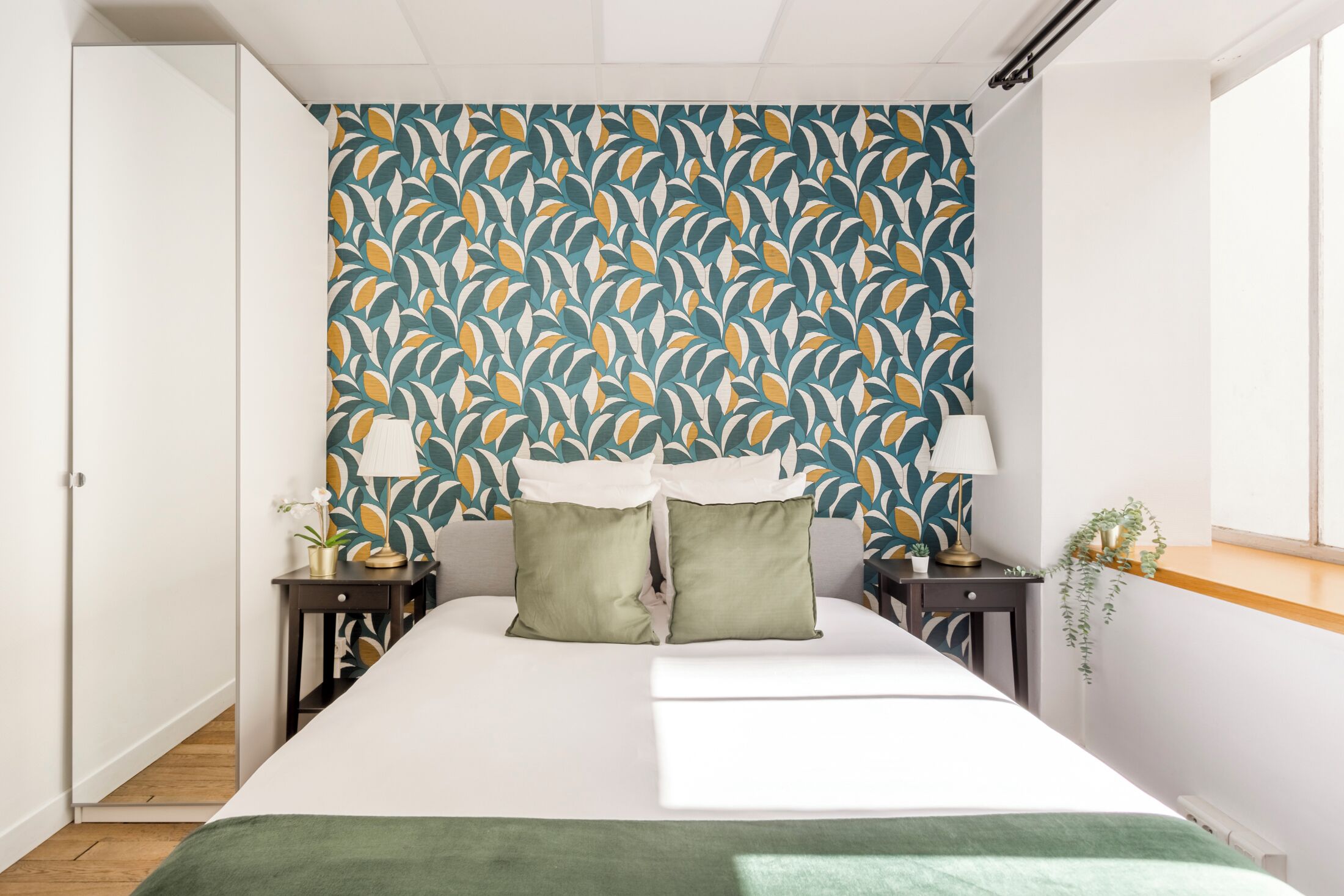 A bedroom with a patterned wallpaper, a bed with green pillows, two bedside tables with lamps, and a potted plant.