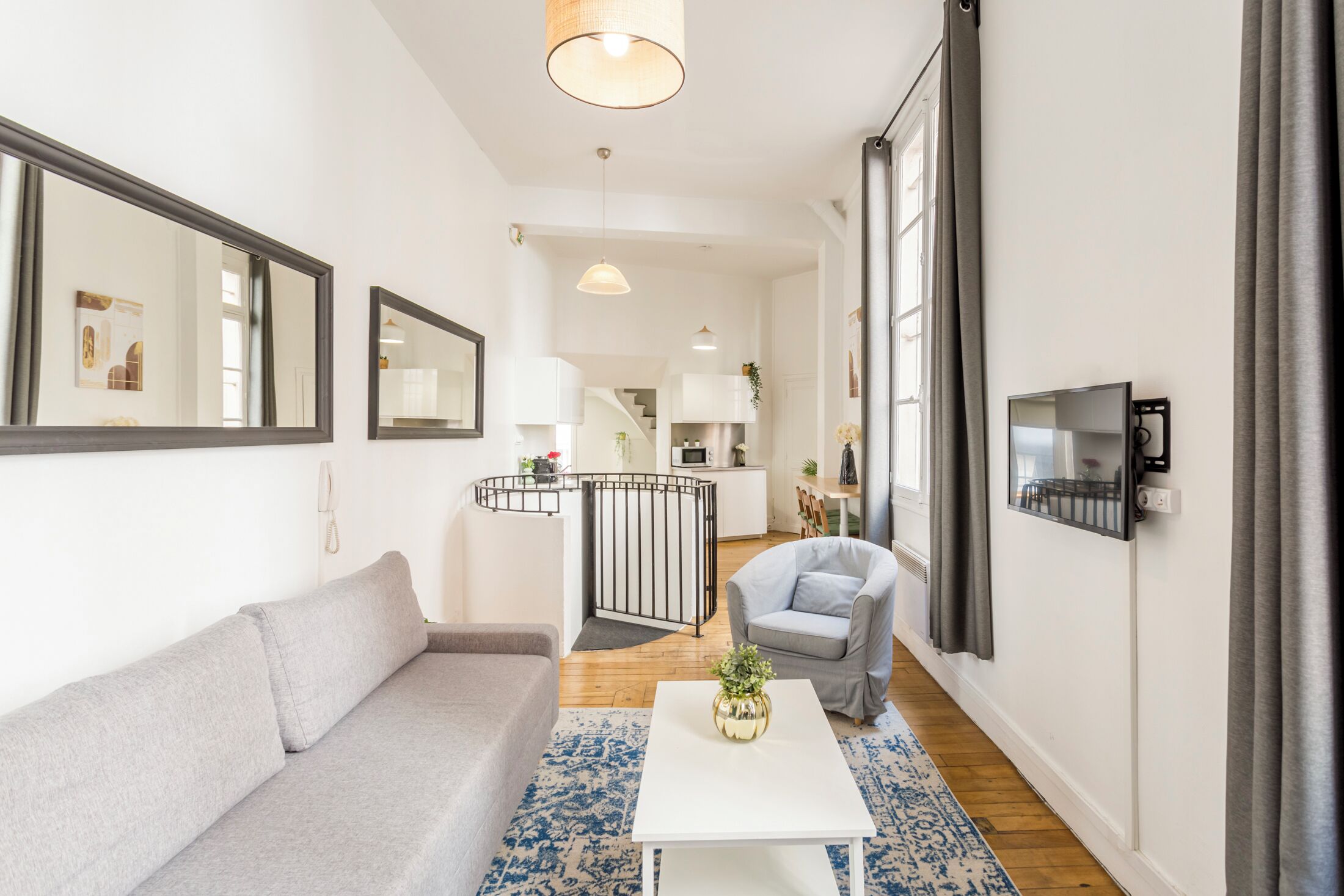 A modern living room with a grey sofa, a white coffee table, and a grey armchair. There is a kitchen area in the background with a staircase leading to an upper floor.