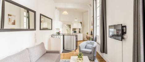 A modern living room with a grey sofa, a white coffee table, and a grey armchair. There is a kitchen area in the background with a staircase leading to an upper floor.