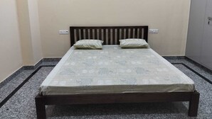 Una cama individual con cabecera de madera, dos almohadas y una colcha estampada en una habitación con paredes lisas y suelo de baldosas.