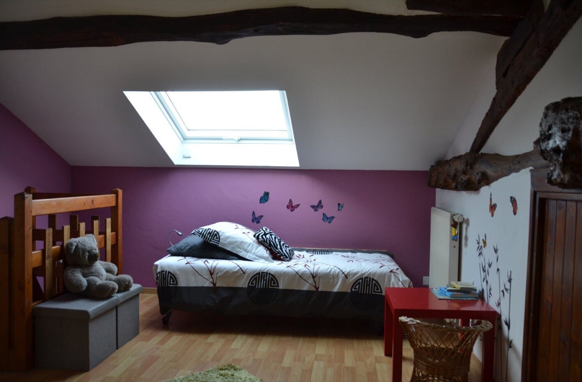 Ein Schlafzimmer mit schrägem Dach, einem Oberlicht, einem Bett mit gemusterten Betttextilien, einem Holzstuhl, einem kleinen Tisch und einer Wandverzierung.