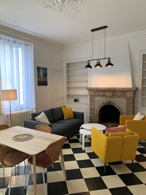 Sala de estar com lareira, piso xadrez e área de refeições com cadeiras de vime e uma mesa.