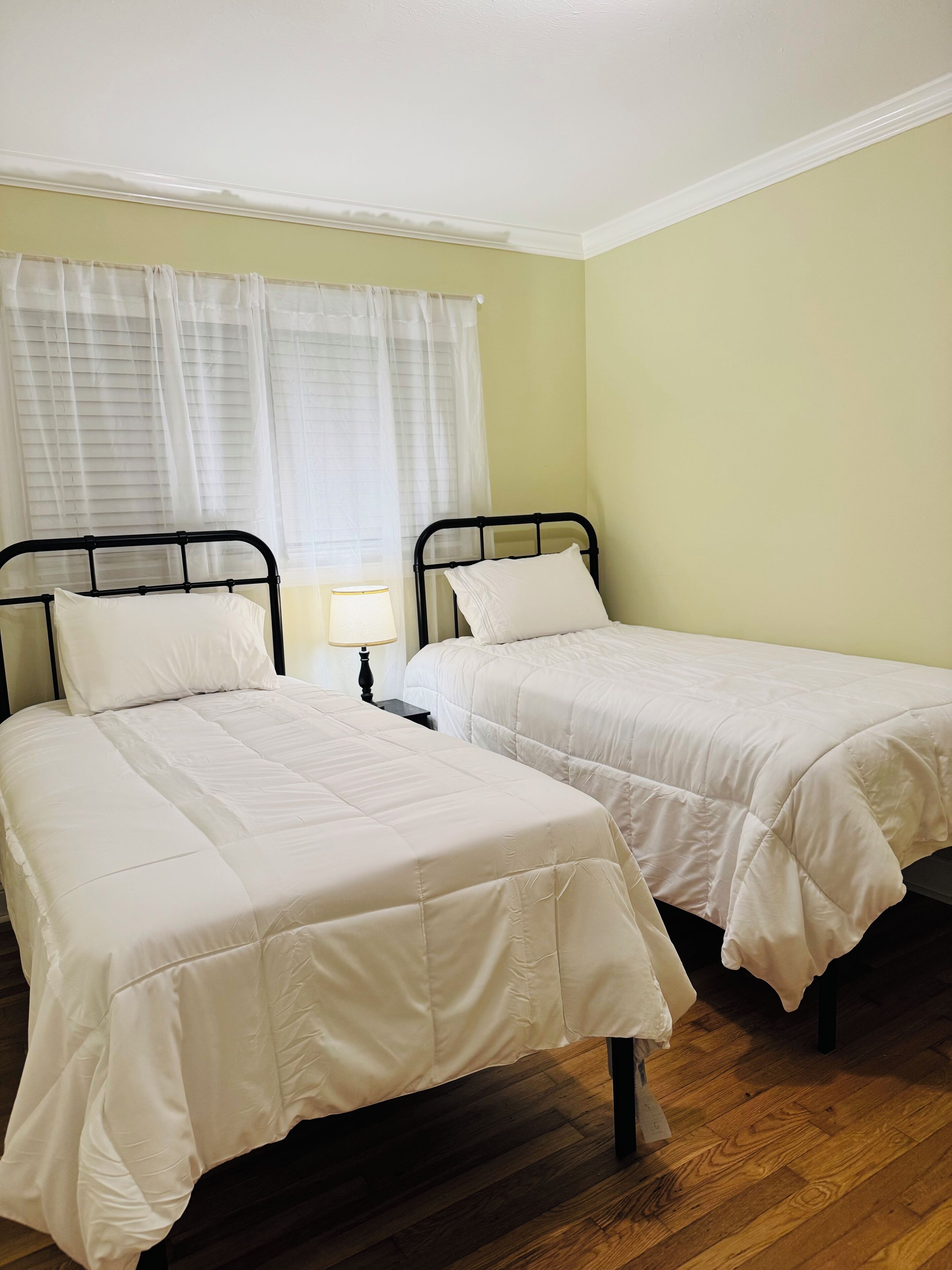 Une chambre avec deux lits simples, une table de chevet avec une lampe et une fenêtre avec des rideaux légers.