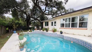 Una piscina con bordo piastrellato, circondata da vasi con piante e una casa con ampie finestre.