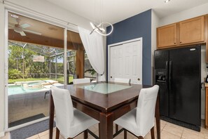 Un coin repas avec une table à plateau de verre, des chaises blanches, un réfrigérateur noir et une vue sur une piscine, visible à travers des portes vitrées.