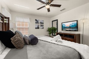 Une chambre à coucher avec un lit, des oreillers, un ventilateur de plafond, une télévision et des œuvres d’art murales.