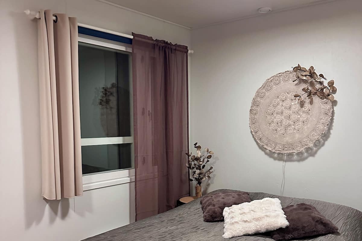 Une chambre à coucher avec un lit, une fenêtre avec des rideaux, une assiette décorative accrochée au mur et un petit vase contenant des fleurs séchées.
