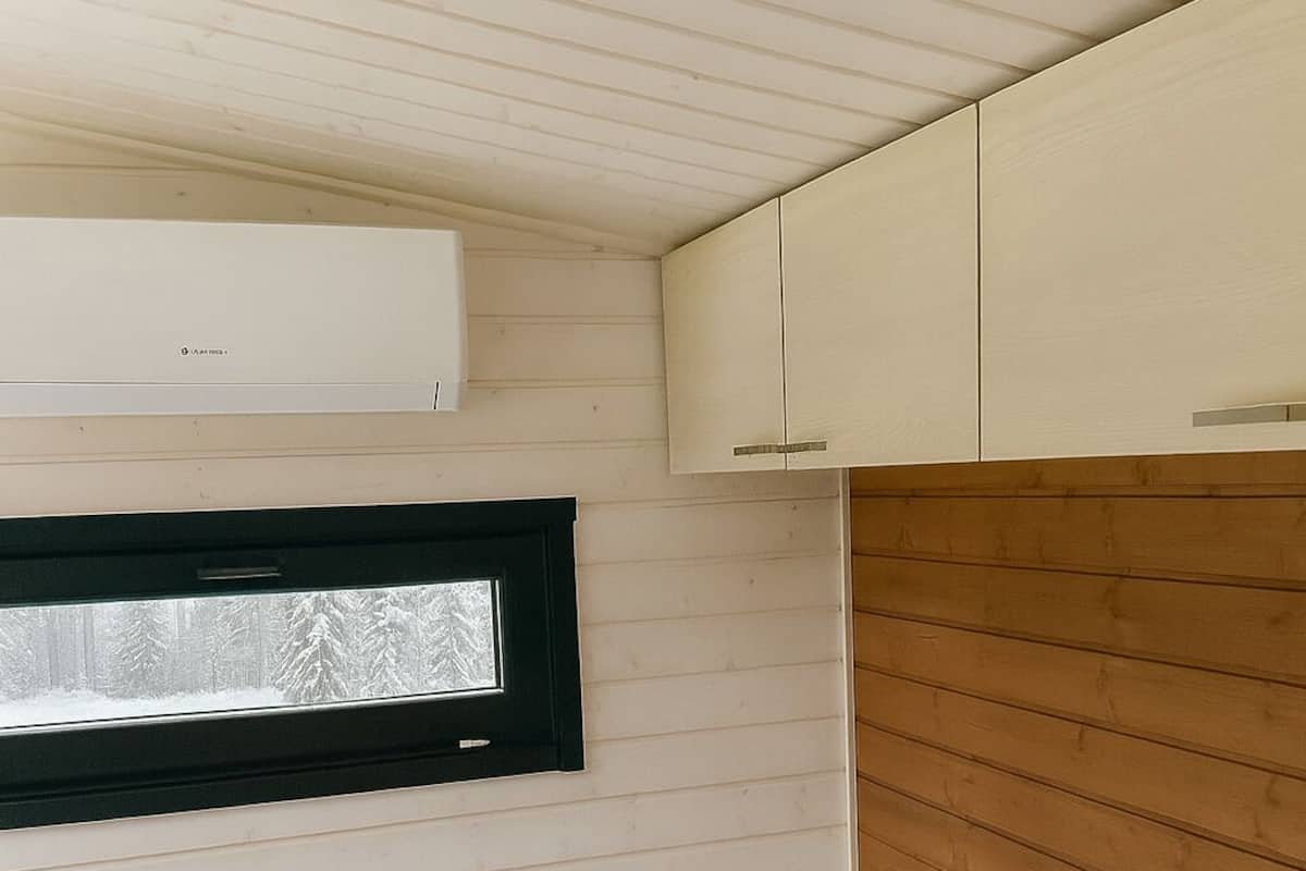 Une pièce compacte et bien éclairée, avec un lit, une fenêtre donnant sur des arbres couverts de neige, un appareil de climatisation blanc et un lambris en bois sur les murs.
