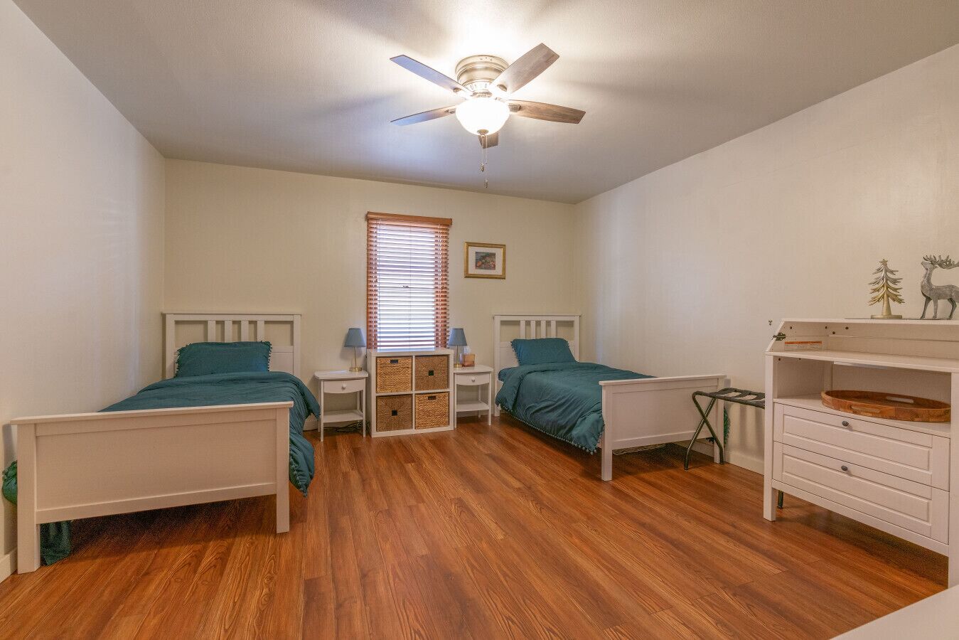 Une chambre avec deux lits simples, un ventilateur de plafond, une fenêtre avec des stores, une table de chevet avec une lampe et une commode avec un miroir.