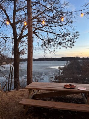 Une table de pique-nique en bois, avec une assiette de nourriture, un banc et une vue sur un lac gelé, bordé d’arbres et illuminé de guirlandes lumineuses.