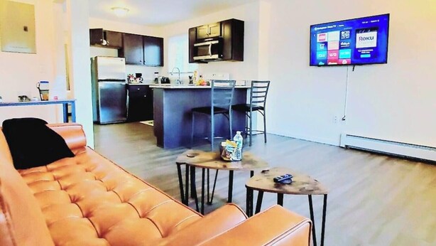 Uma sala de estar moderna com um sofá de couro, uma área de jantar com uma mesa pequena e cadeiras, uma cozinha com armários escuros e uma geladeira, e uma televisão de tela plana presa na parede.
