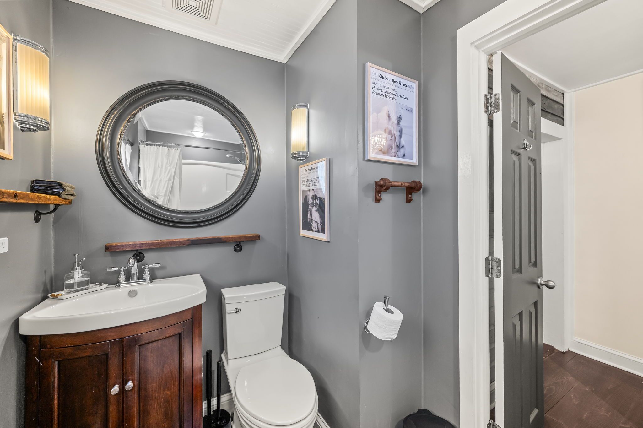 A bathroom with a round mirror, a white sink, and a toilet. There are framed pictures on the wall and a wooden shelf above the sink.
