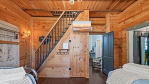 A wooden cabin interior with a staircase leading to a loft bedroom, a bed with a white comforter, and a wooden wall with a picture.