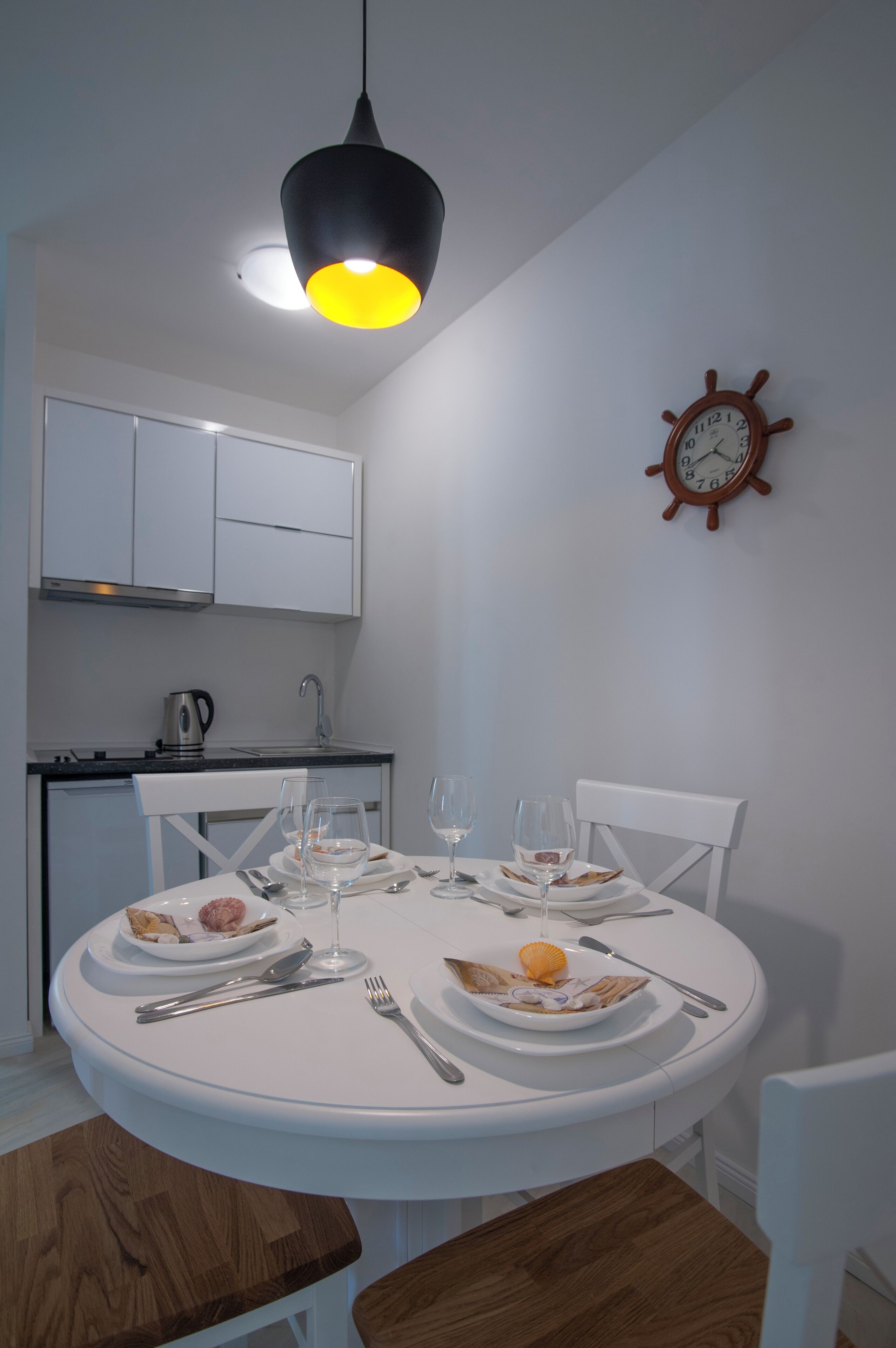 Un comedor con una mesa redonda dispuesta para dos personas, sillas blancas, un banco de madera, una cocina al fondo y un reloj de pared.