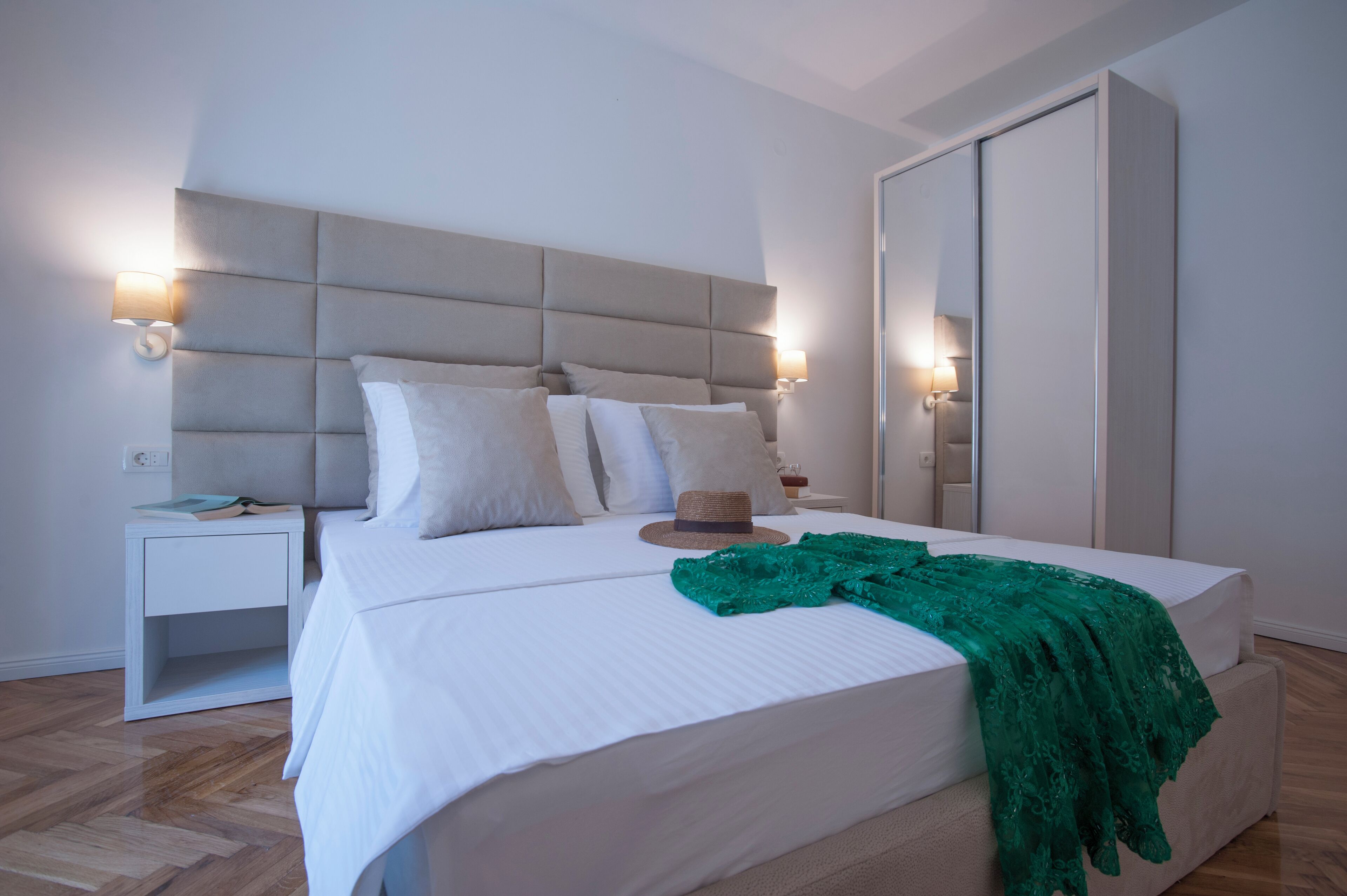 Una cama bien tendida con un cubrelecho de encaje verde, una lámpara de noche y un sombrero en la mesita de noche.