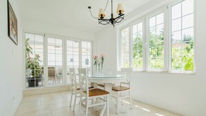 A bright dining area with a glass table surrounded by white chairs, a chandelier, and large windows with a view of greenery outside. - Captain's Villa (Budva)