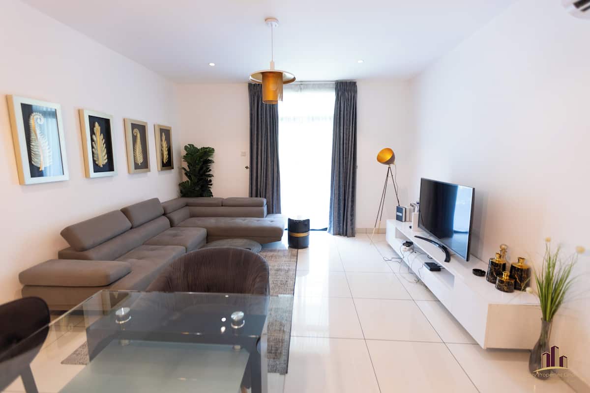 A modern living room with a sectional sofa, a glass dining table, a flat-screen TV, and decorative items on a white console.