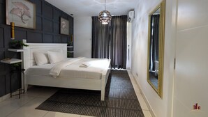A modern bedroom with a white bed, a geometric pendant light, a large mirror, and a framed floral artwork on the wall. - RoyalRest at Embassy Gardens (Accra)
