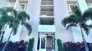 Un hotel moderno con una entrada central, varios balcones y un paseo empedrado con plantas.