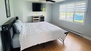 Une chambre à coucher avec un lit, un ventilateur de plafond, un téléviseur fixé au mur, une fenêtre avec des stores et un plancher en bois.