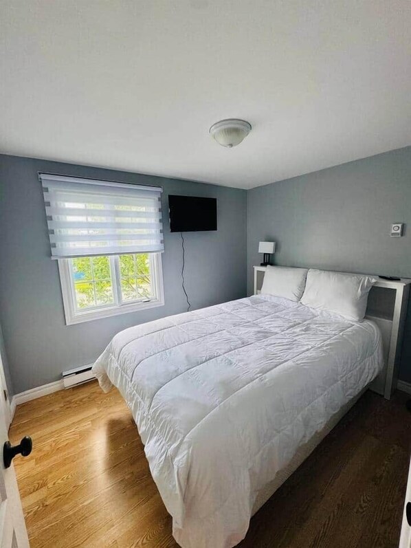Een slaapkamer met een bed, een raam met jaloezieën, een wandtelevisie en een plafondlamp.