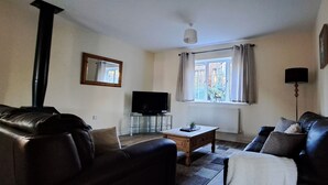 A living room with a leather sofa, a coffee table, a flat-screen TV on a glass stand, a window with curtains, and a radiator. - Sizeable 4 bed property on Huntingdon Ring road (Huntingdon)