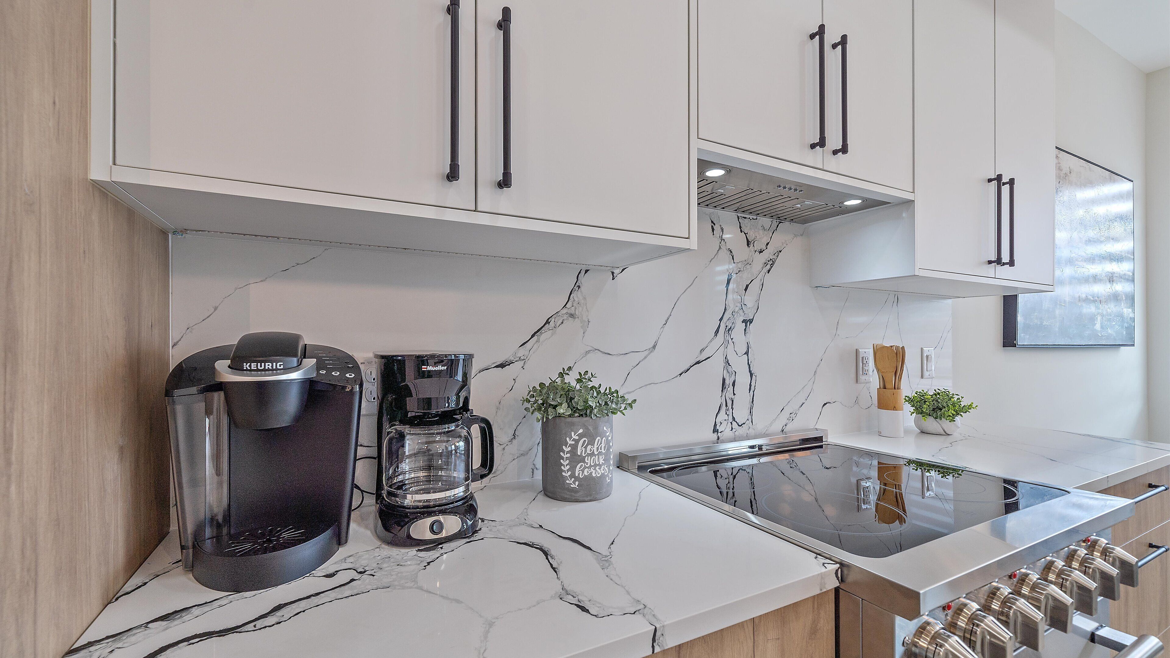 Una cucina moderna con mobili bianchi, una macchina da caffè nera, un piano di lavoro in marmo e una cappa aspirante in acciaio inossidabile.