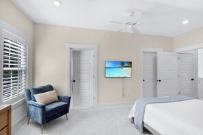 Une chambre avec un lit, une chaise, un ventilateur de plafond et un téléviseur fixé au mur.